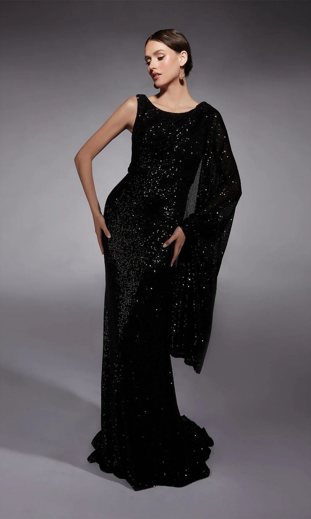 A woman wears the Alyce 27718 long formal black dress, sleeveless and adorned with sequins, paired with a draped shawl, standing against a plain gray background.